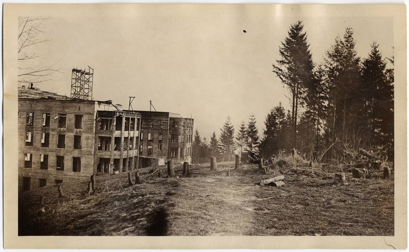 Sepia tone photograph of Multnomah County Hospital, showing a large cleared field with evergreen trees in the distance, and a half-constructed four-story building in the foreground.