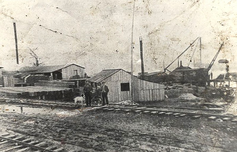 A black and white photo of an old factory with workers standing beside it.