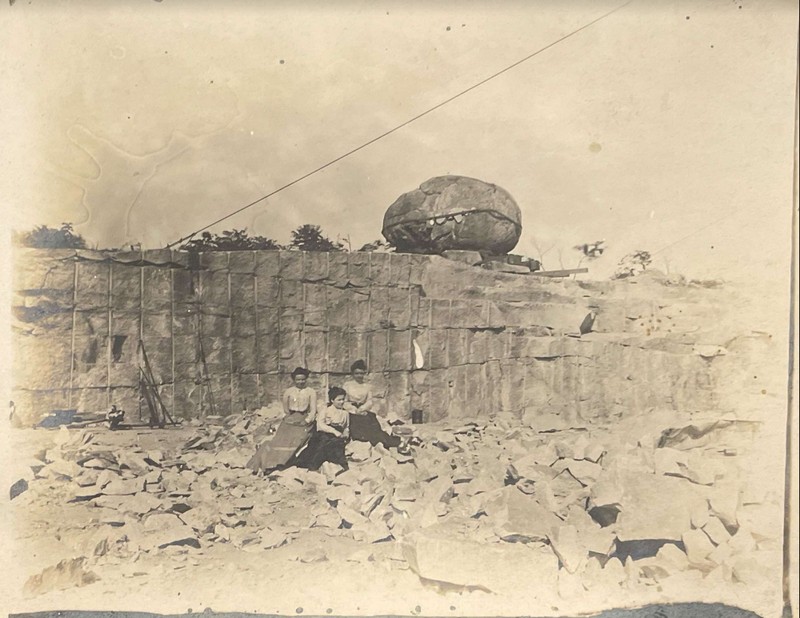 A black and white photograph with three people sitting in center of frame, with the Rollstone boulder in the background above them.  