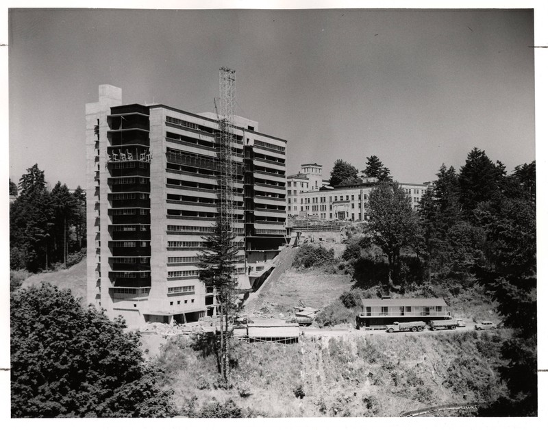 A partially constructed, tall, midcentury hospital building is visible on the side of a hill. Campus buildings are visible in the background.