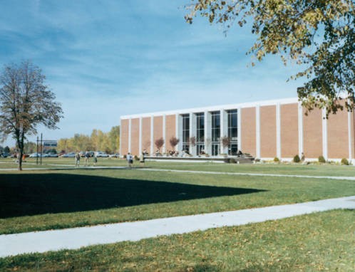 Color photograph of the front of the library as seen from near the Student Union, ca. 1966. Below the tree on the left is a sign the reads "Hello Walk." 