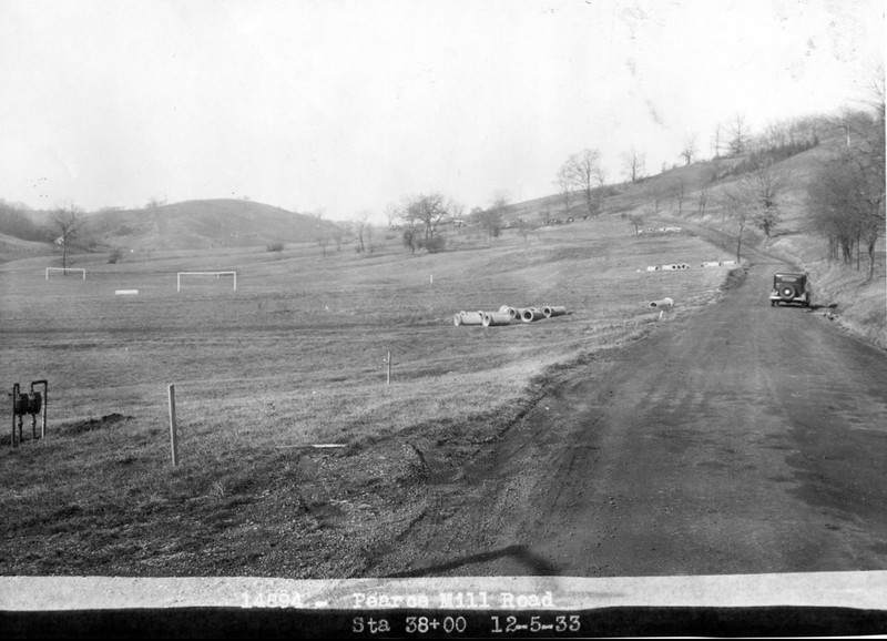 Black and white image showing a dirt road extending into the background. A single car is on the road in the distance, and large pieces of pipe lay in the field on the left side.