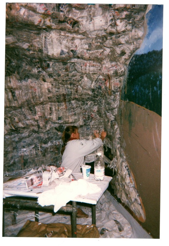 The artist sits on the floor in the center of the image, applying the papier mache over the foamcore to create the shape of the rock shelter. She sits next to a small table that holds the paper mache materials. 