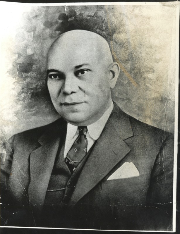 A portrait of a middle-aged African-American man. He is bald and clean-shaven, and is looking at the camera with a neutral expression. He is wearing a dark-colored suit and vest with a white shirt and a dark-colored tie. A small tie chain is visible around his tie, and he has a light-colored pocket square.