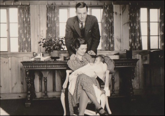 Black and white photograph of a young man in a suit and tie leaning over the chair with his arms on his wife's shoulders. The young woman in a dress in the chair is cradling an infant wrapped in a blanket. In the background is a long wooden table with ornamental legs and windows cover the walls.