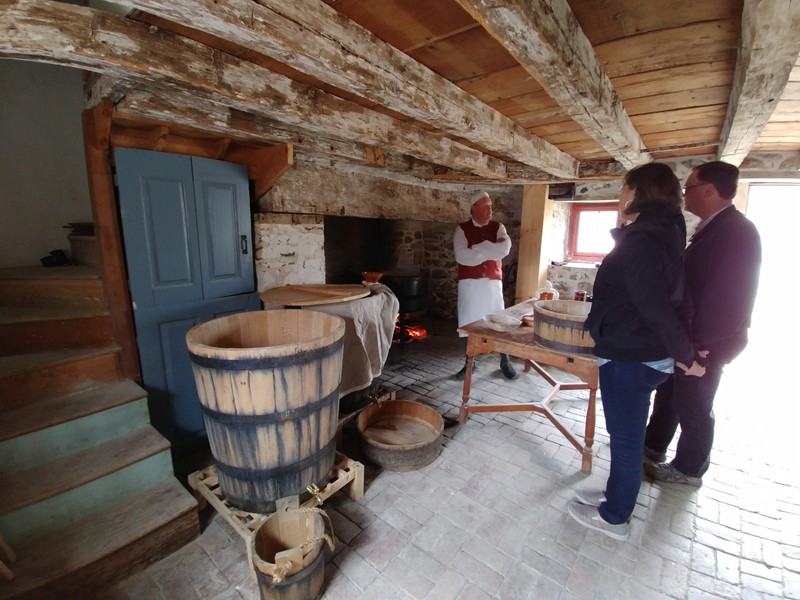 Image of man in colonial clothing standing in front of a large fireplace and talking to 2 visitors across a work table.