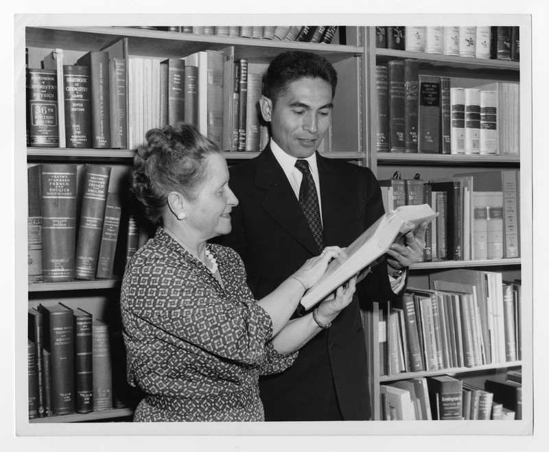 Black and white photograph of Bertha Hallam, in a floral patterned dress, assisting a student in a suit, in front of library bookshelves.