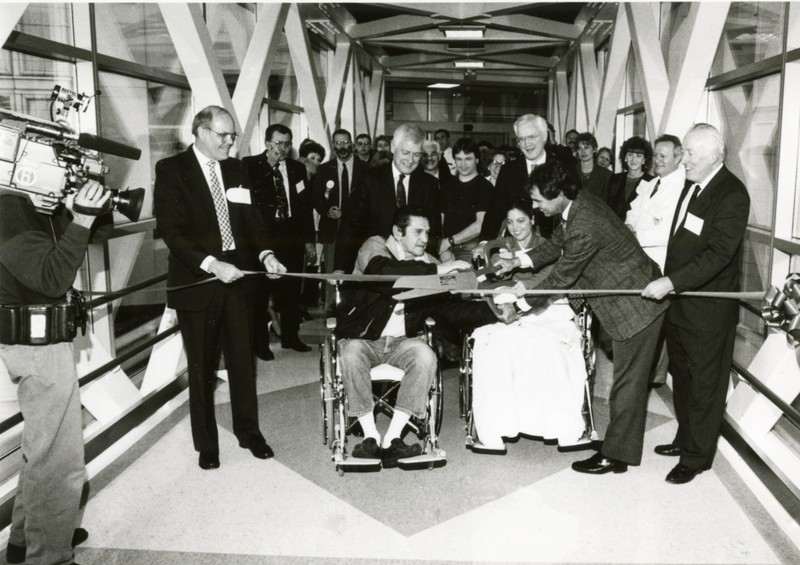 A crowd of onlookers watch as four standing people and two people using wheelchairs use large novelty scissors to cut a ribbon on the glass and metal pedestrian sky bridge.