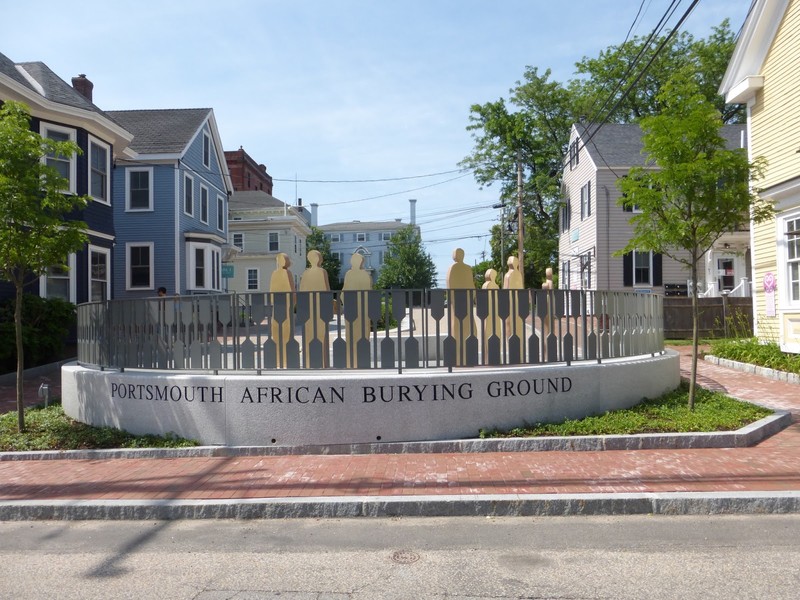 A sequence of figures representing the community of Portsmouth uniting and honoring the memory of the burial grounds and form a ring around the burial vault that displays the West African Sankofa symbol.