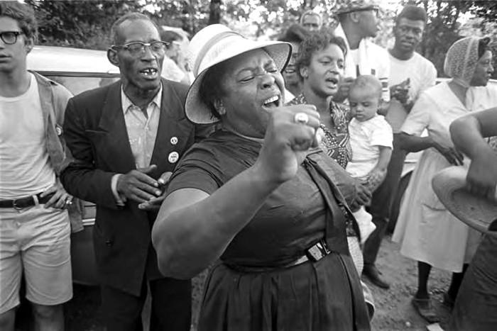 A half-length photo of Hamer in a light hat and dark dress. She is singing, right arm upraised, with about a dozen March participants standing behind her.