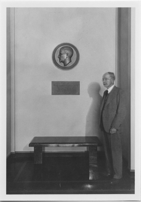 Black and white photograph of John E. Weeks, M.D., standing next to the dedication plaque and bench in the foyer of the Library and Auditorium. The plaque is in honor of Dr. Weeks who donated $100,000 towards the construction of the Library.