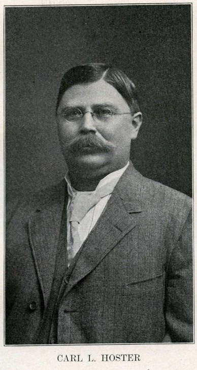 Carl L. sitting, looking forward while wearing a suit and a tie with glasses and a mustache