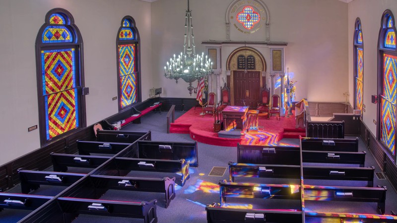 Mahagony pews with red cushions surrond the Bimah, a raised platform, at the front of the synagogue. The Bimah is backed by the flag of Israel and a stained glass window.