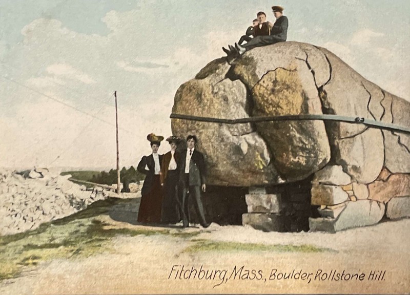 A colored photograph of 6 people posing with the rollstone boulder, 3 people on top of it sitting, 3 in front of it standing.  The Boulder has a brace on it to keep it together and is connected to more pieces of rock on the right side of the image.  