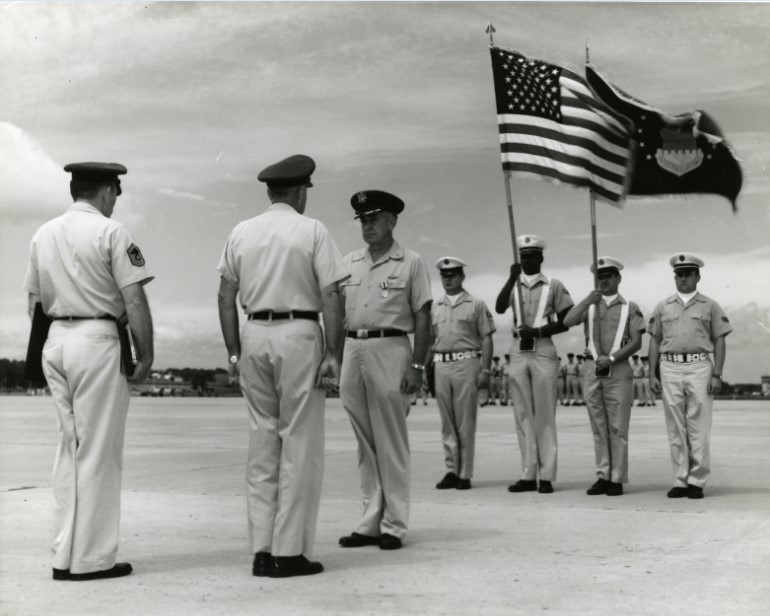 Photograph of a ceremony at Truax Field, featuring Colonel Mace and a Color Guard. Featured in the background of the image are other Air Force personnel and Truax Field.  