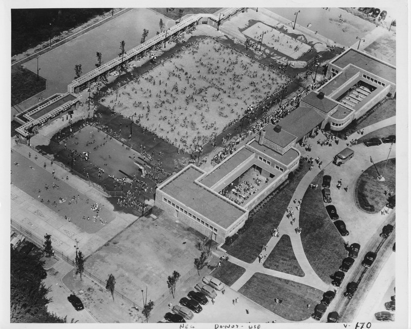 A grayscale aerial photograph of a crowded pool complex. There is a large central pool, with two smaller pools on either end. All three pools are full of people, with additional crowds on the pool deck. On the right side of the image, a building can be seen, housing the entrance and open-air locker room areas.