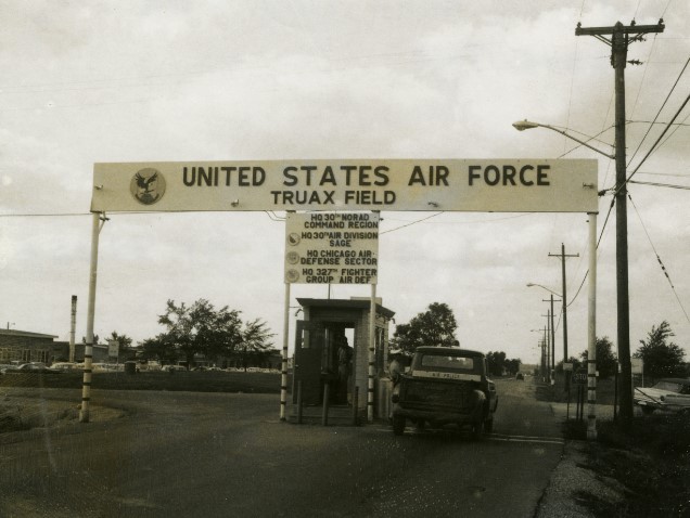 Image of the main gate at Truax Field, taken during the Cold War. Above a guardhouse is a sign that reads “United States Air Force Truax Field” with a list of units based at the field below the main sign. In the image, a vehicle is marked “Air Police” with buildings and cars beyond the gate. 