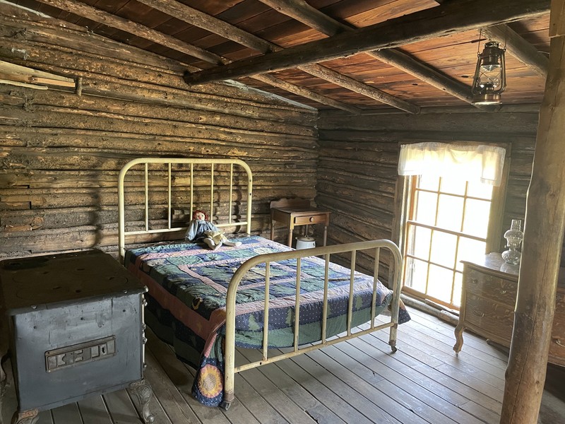 A photo of a log cabin bedroom showing a stove, large bed, and bedside table with a chamber pot underneath. There are two small dolls at the head of the bed.