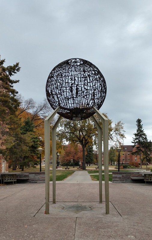 Metal mesh globe surrounds eternal flame. Held up by metal supports. Stands on open plaza.