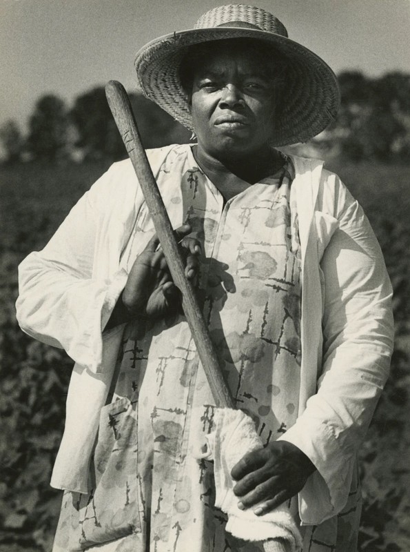 Fannie Lou Hamer, wearing a workdress, jacket, and straw hat, faces the camera in a 3/4 length portrait. She is standing in a field and grasping the wooden shaft of a tool, perhaps a shovel or hoe.