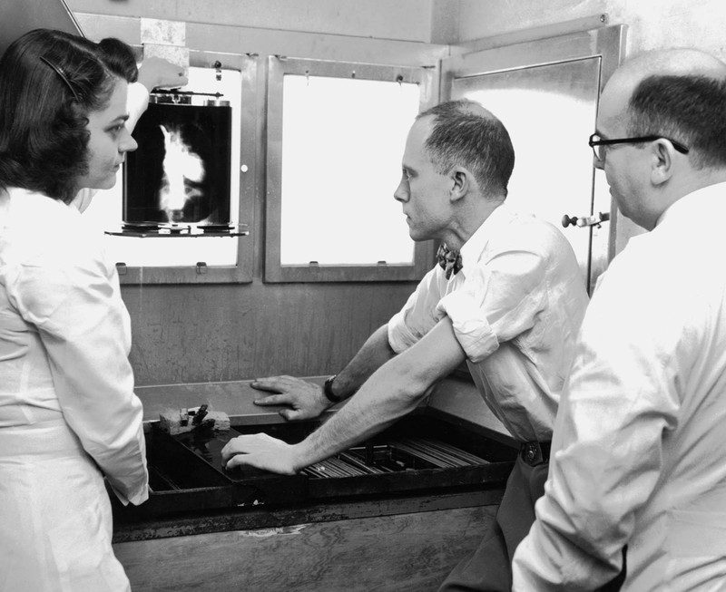 Black and white photograph depicts Miss Kay Smith (left), x-ray technician, who holds up x-ray made of patient's heart who has a congenital heart condition. Dr. Charles T. Dotter, radiologist (center) and Dr. Herbert Griswold, heart specialist (right), make diagnosis from the film so that they can recommend to surgeon exact spot where heart repair must be made.