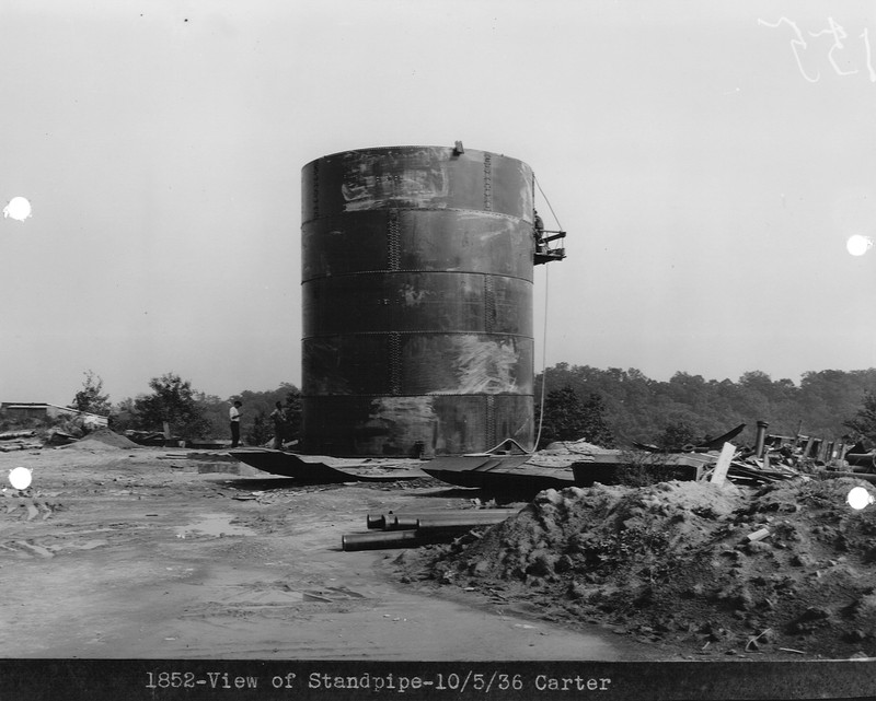 Black and white image shows a large round metal structure, constructed from five vertical rows of large panels stacked on top of each other. Each panel is as tall as the two people standing at its base, which has a platform attached by ropes to its side with a third person standing on it. Construction debris, such as pipes, metal sheets, and mounds of dirt are scattered nearby in the foreground.