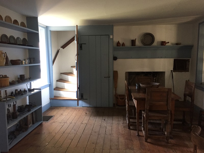 Image of a room with an enclosed set of stairs on the left side; a wooden table and chairs to the right, with a fireplace in the background.