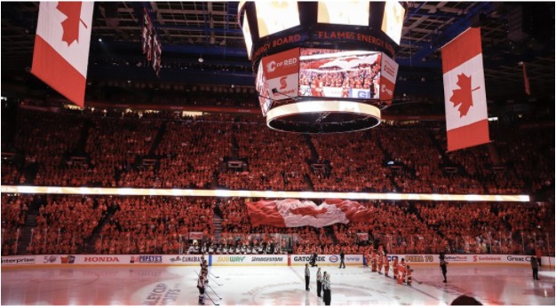 This image was captured inside the Saddledome right before one of Canada’s most significant sporting events: an NHL game for the Calgary Flames. The image is taken from the perspective of a fan in the audience cheering for the Calgary Flames during a game. The players, coaches, refs and fans are all standing, facing the Canadian flag while the national anthem is being sung. This image encompases Canadian identity and nationalism through its red colours, Canadian flags being passed through the crowd, collective admiration for the game of hockey and paying their respects to the country. The stadium appears to be sold out with the majority of the fans wearing Calgary jerseys. There is a sense of pride and community within the stadium as tens of thousands of strangers share one thing in common, their love for the Calgary Flames and the game of hockey. Once these strangers enter the Saddledome, they are united as they cheer, “boo” and heckle together. No matter what one may be going through, when that red goal light goes off for their team, one thing is certain, nothing else in the world matters. Tony Bennett’s “The Exhibitionary Complex” explains the feelings behind this image by explaining that people respect certain dress codes and people are happy about the role they are assigned to (lecture expo 67). The people who are cheering become part of the spectacle themselves. Seeing as most of the fans are wearing Calgary Flames memorabilia or the team's colours, it demonstrates a sense of patriotism to support the team.