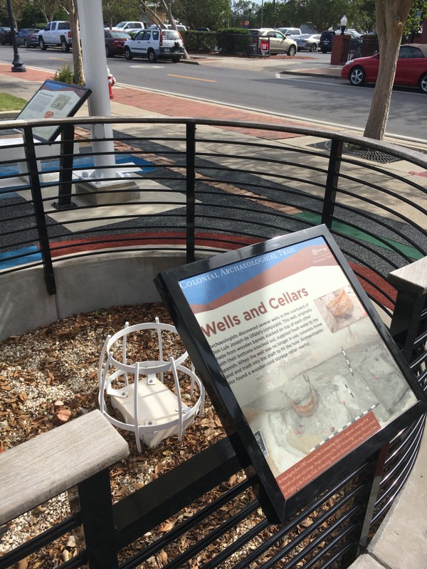 Interpretive "Wells and Cellars" sign on fence. Small pit within fence contains historic brick foundation and metal barrel. 