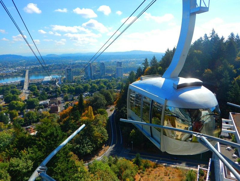 Color photograph depicts small metal tram cabin with many trees in near background and tall buildings and river valley in background.