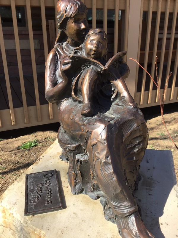 Made of bronze, this small sculpture depicts a woman sitting in a chair with a small child on her lap. They are relaxing in the chair while reading a book. 