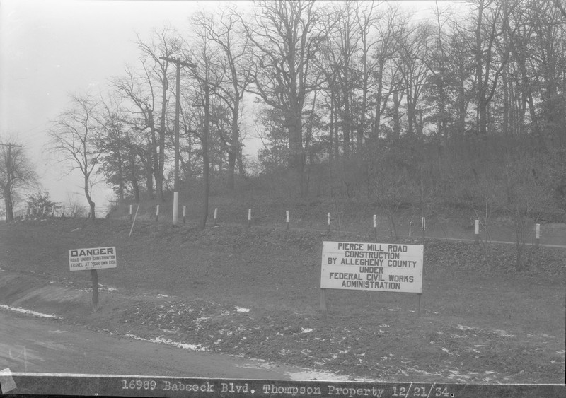 A black and white photo, taken from a road, with another road above it lined with trees. A sign beside the lower road reads "Pierce Mill Road Construction By Allegheny County Under Federal Civil Works Administration."