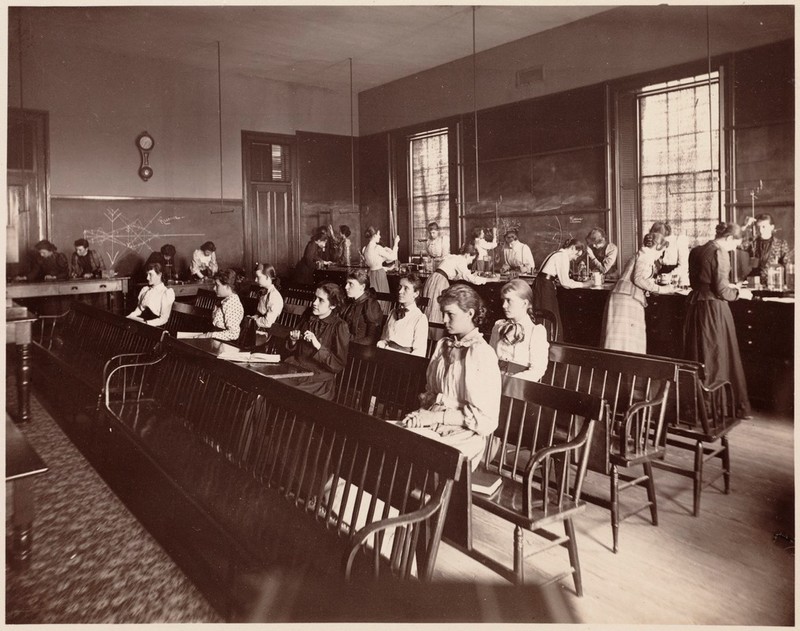 Black & White photo of young women in long-sleeved, long dresses sitting on rows of benches. Background young women standing at lab benches for a physics lab.