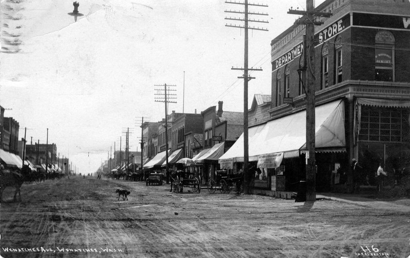 View looking south on Wenatchee Avenue from the intersection with First Street. The Griggs Building with the Wenatchee Department Store on the southwest corner. Many horse drawn wagons and buggies on the dirt roadways. People walking along concrete sidewalks. 