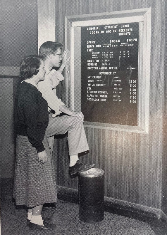 A young man and woman stand in front of a letterboard with events listed