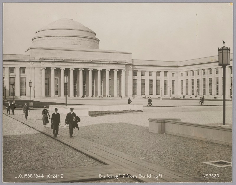 Black & white, domed building with columns across the facade. Gravel ground cover with wooden plank walkways. People walking.