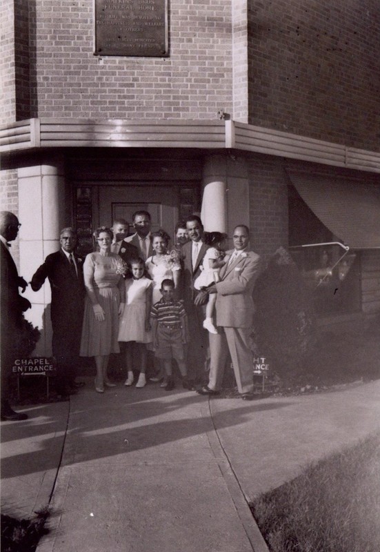 The image shows a group of twelve people (Six men, three women, two young girls, and a young boy) standing in front of the front door of a building. Some distance above the door, the bottom of a metal plaque is visible. Text on the plaque reads "THERON B. WATKINS  Founder  WATKINS BROS FUNERAL HOME  His Life Was Dedicated To The Happiness And Welfare Of Others.  This Tablet Dedicated By His Many Friends".