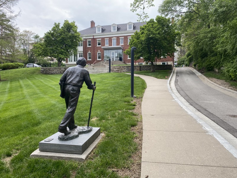 A statue of Benton. He is holding a sketch book and waling with a walking stick. A large brick building is in the background.