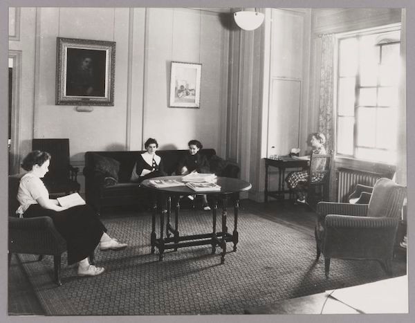 Black & White photo. Formal seating area. Two women sitting on a sofa talking and reading magazines.