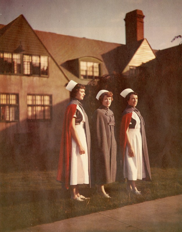 Three young women stand in white aprons, caps, and blue and red capes in front of a large storybook style building lit by the sun.