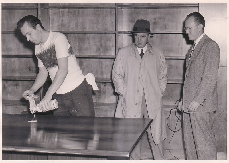 Three men stand in a wood-paneled office with a desk in the foreground. To the left, a man oils the wood on the desk.