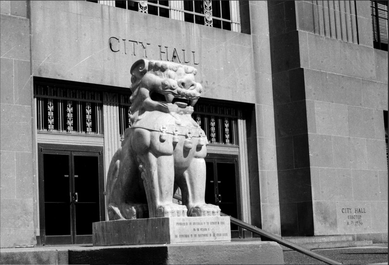 An image of one of the lion statues that sits on the South Plaza steps. The base of the statue features the English version of the inscription reading: "Presented by the government of the Republic of China on the occasion of the Bicentennial of the independence of the United States." In the background can be seen the South entrance to City Hall and the cornerstone of the building bearing the inscription, "City Hall, A.D. 1936."