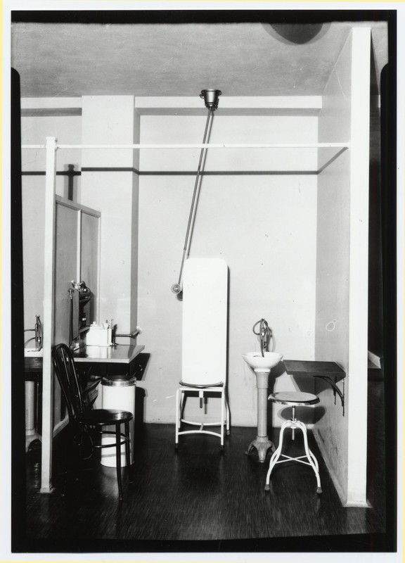 Black and white photograph of the Outpatient Clinic examination area at the University of Oregon Medical School, with a chair, foldable patient bed, small sink, and other supplies.