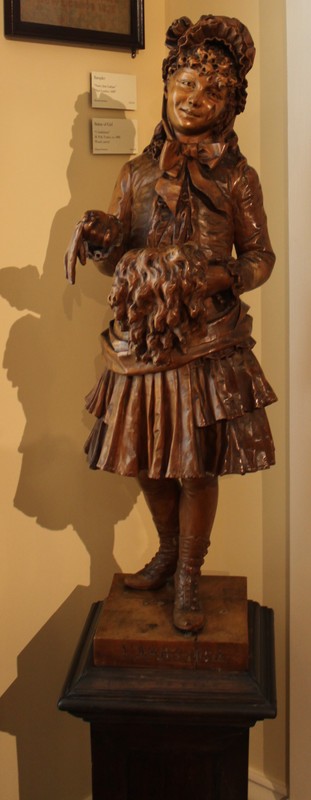 Carved Wood Statue (Italian, 19th Century). The statue is of a young girl in a dress, bonnet and boots holding a fur muff and a glove. The statue is signed on the front of the wood platform base, "L'Ambiziosa" and it is signed on the side of the base, "M. Poli, Venezia".