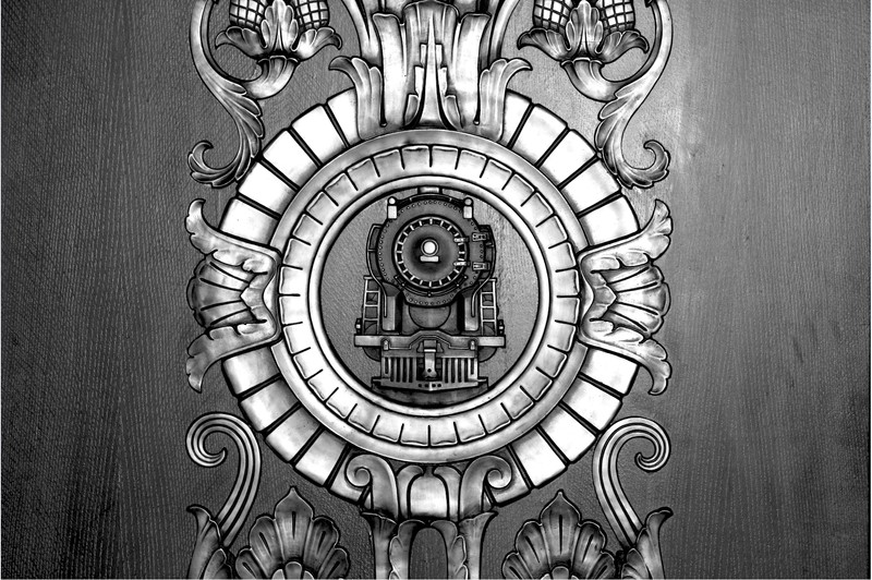 A close-up, black-and-white, detail image of the bronze metalwork that displays a train surrounded by two segmented concentric circles and scrollwork expanding symmetrically in four directions.