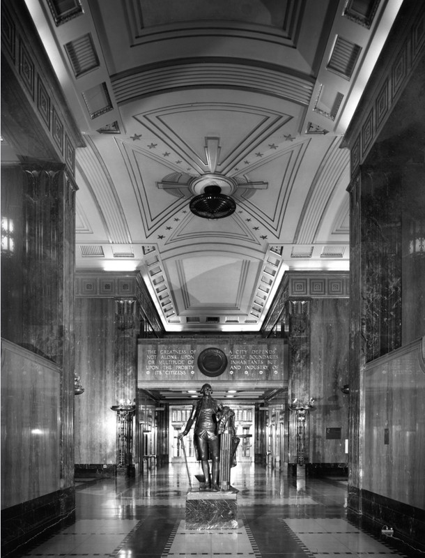 A black-and-white photograph of City Hall's Main Lobby and Rotunda showing a statue of President George Washington with a cane in his right hand and left hand resting on a fasces. Behind and above the statue is the inscription of a quote from Councilman A. N. Gossett reading: "The greatness of a city depends not alone upon great boundaries or a multitude of inhabitants but upon the probity and industry of its citizens."