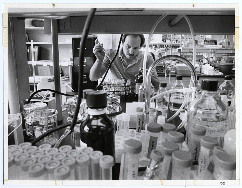 View of a researcher holding a pipette in a laboratory, seen through an open shelf of laboratory equipment.