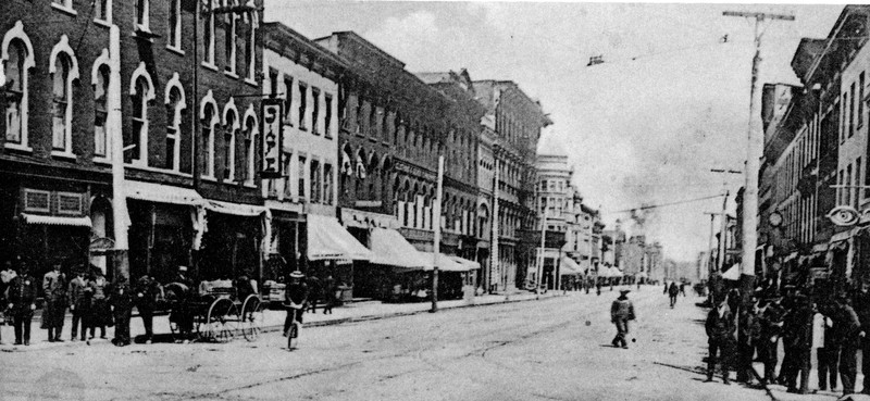 A black and white photo of a street scene at the turn of the 20th Century. A row of building is seen on the opposite side of the street from us. At left are a police officer, a group of men in suits, a carriage, and a woman riding toward the camera on a bicycle. At center and right, men are crossing the street or crowding the sidewalk. 