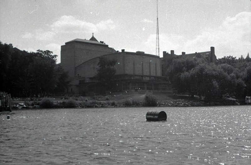 Photograph of Memorial Union taken from Lake Mendota. In the image, a barrell notes the edge of the swimming area, people are gathered on a the lawn, and Memorial Union sits above the lakeshore.  