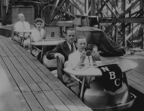 Four men aboard a rollercoaster, two holding recording equipment, with an NBC pennant hanging from the front (black and white) 